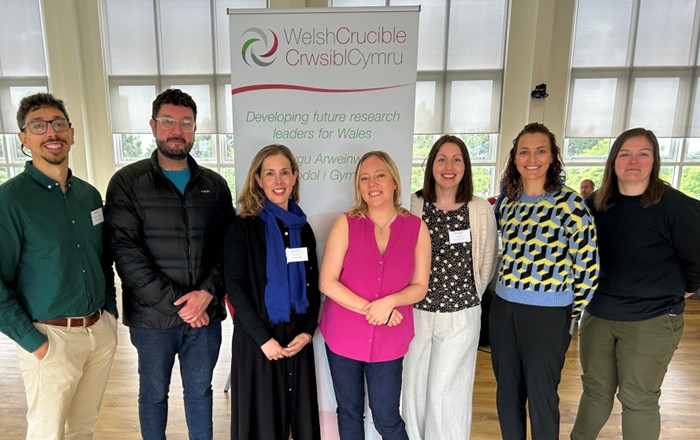 A group photo of the seven Swansea University researchers standing together and smiling in front of a branded pull-up banner displaying the Welsh Crucible logo.