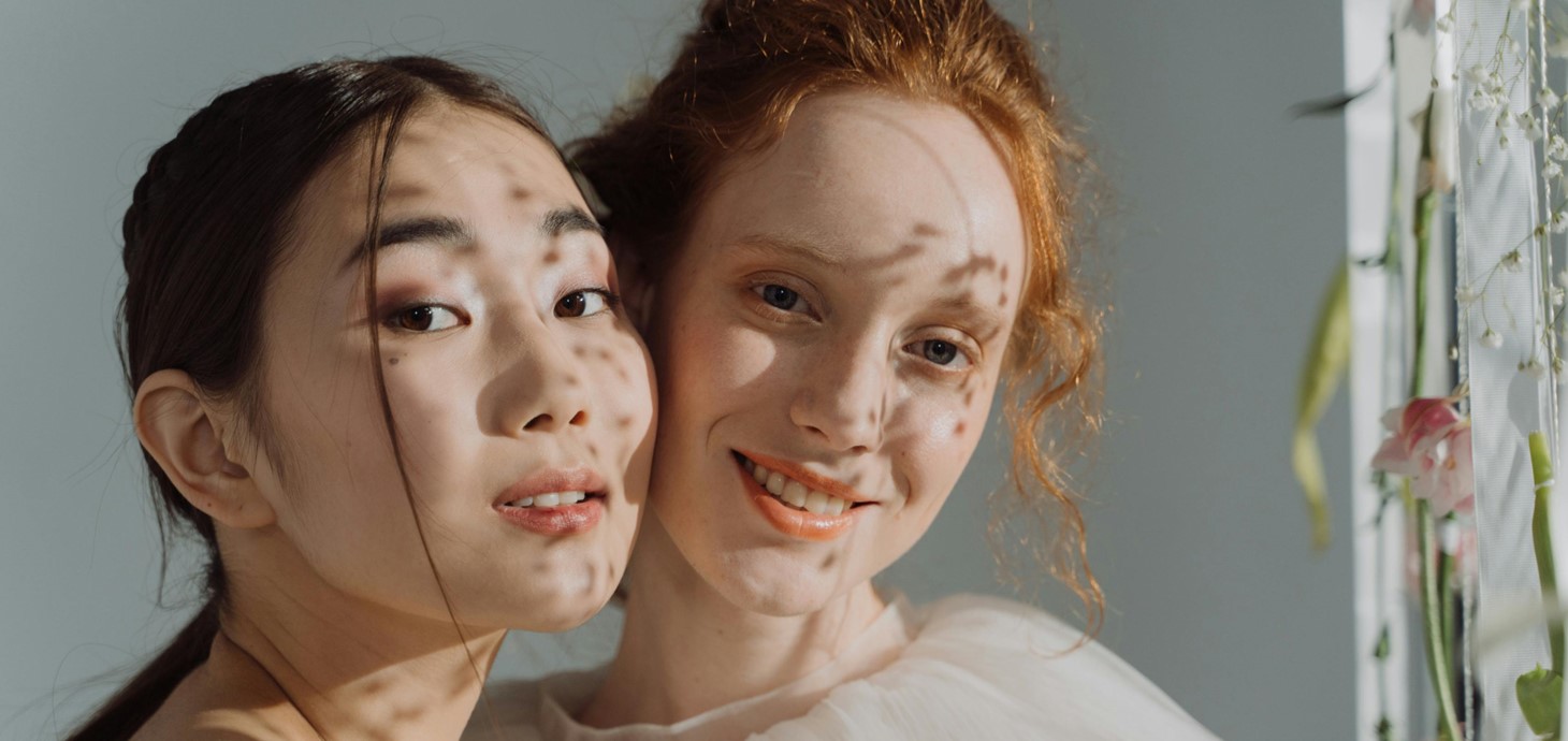 Two young women - one Asian, one Caucasian - standing cheek to cheek looking at camera.
