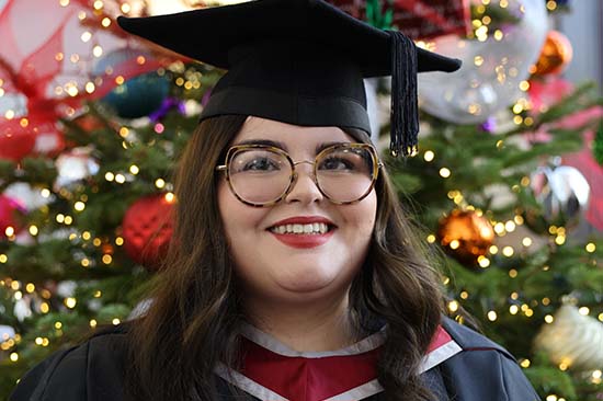 Young woman in graduation robes standing in front of a decorated Christmas tree