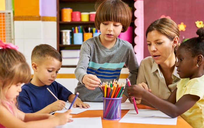 Four young children and one woman sitting round a classroom table using pens and paper