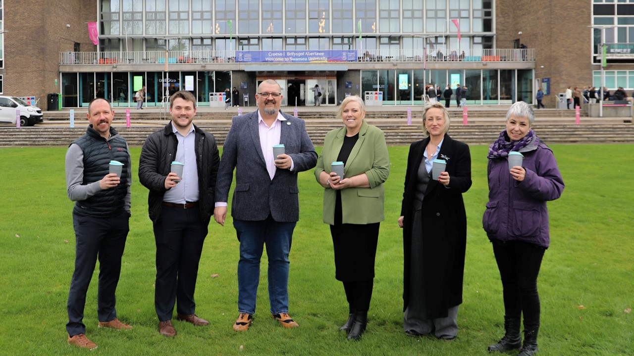 Six people standing on grass in front of a building holding reuseable coffee cups