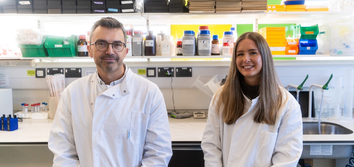 Man and woman wearing white coats standing in front of laboratory equipment.