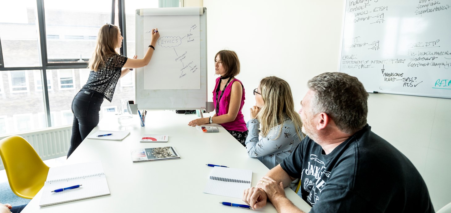 Woman writing on white board in front of three other people in a meeting room.