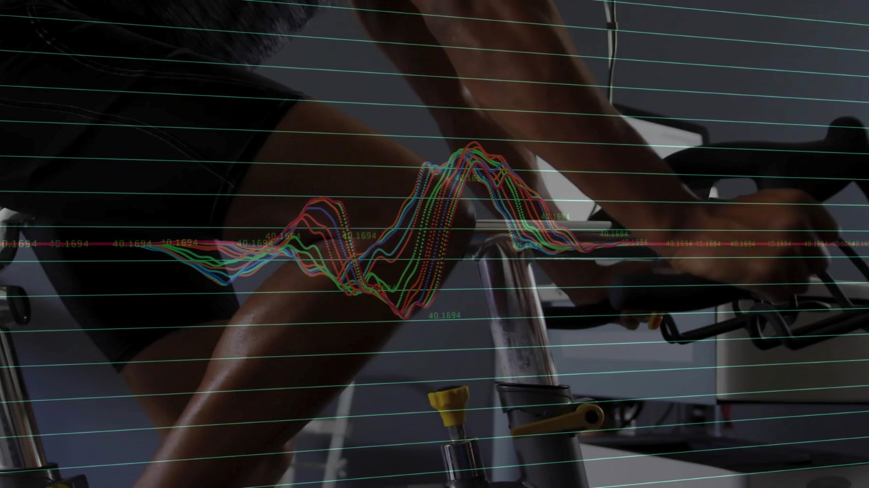 A man rides a stationary bike in a fitness lab as monitoring devices track his performance, shown by an overlaid waveform. Credit: vectorfusionart | Shutterstock.