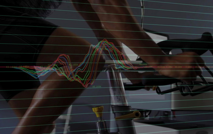 A man rides a stationary bike in a fitness lab as monitoring devices track his performance, shown by an overlaid waveform. Credit: vectorfusionart | Shutterstock.