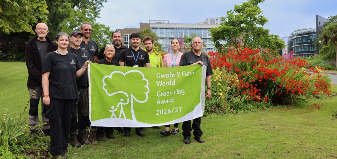 Group of people holding up large green flag in front of a flower border.