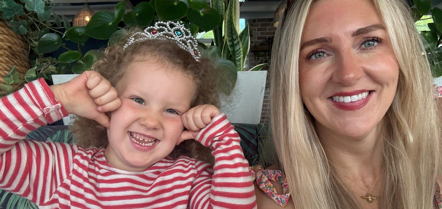 Smiling small girl wearing toy tiara sitting indoors alongside smiling adult woman.