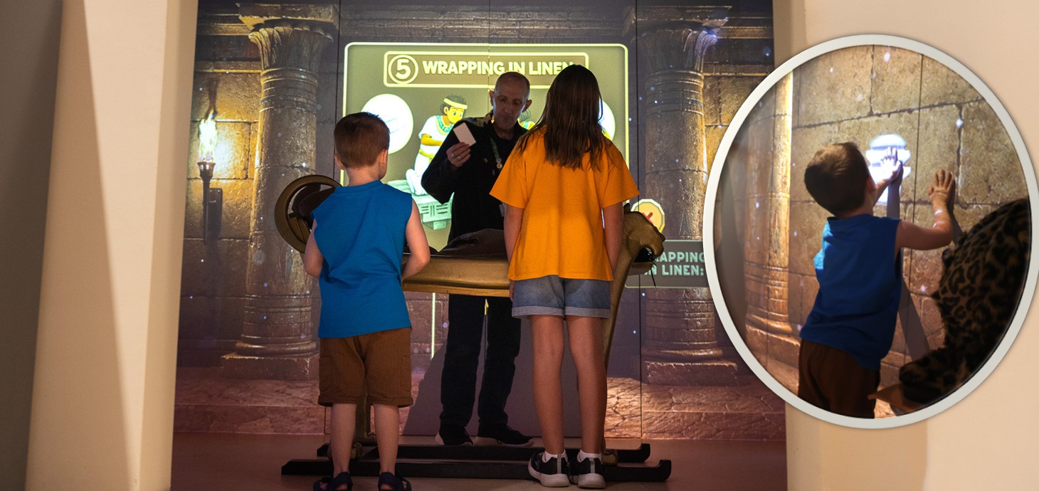 Two children standing in front of a table listening to a man talking with inset picture of a small boy touching a bright area on an interior wall.