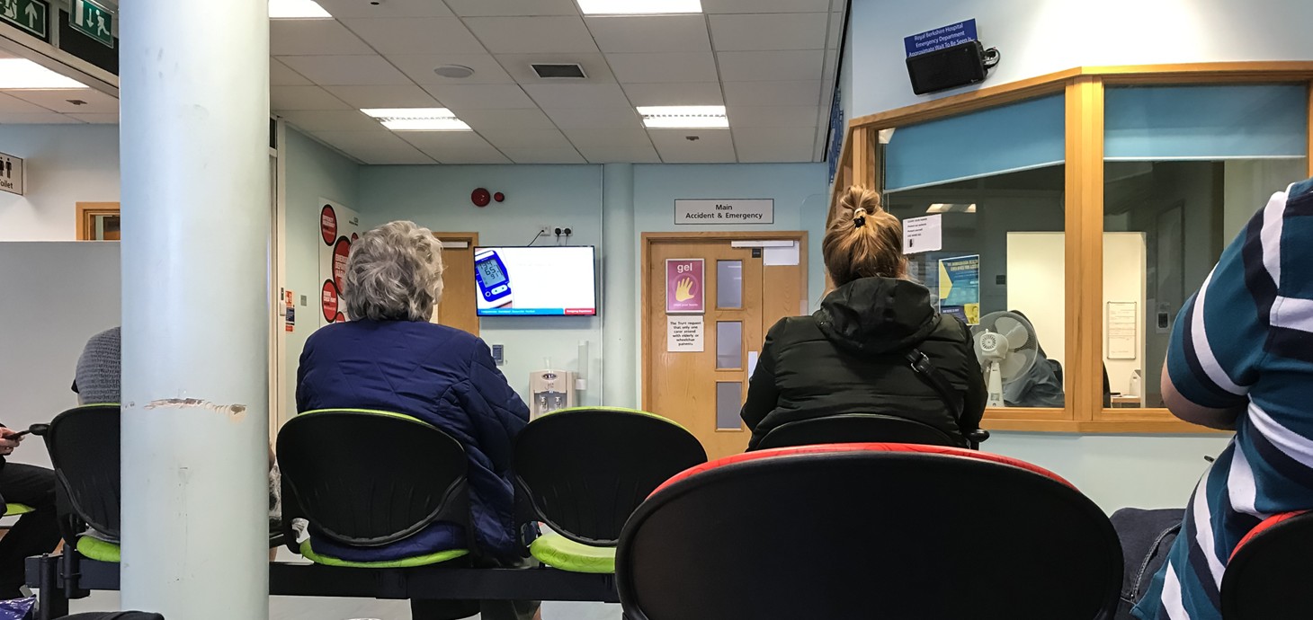 People sitting in a hospital waiting room with a reception desk in the corner.