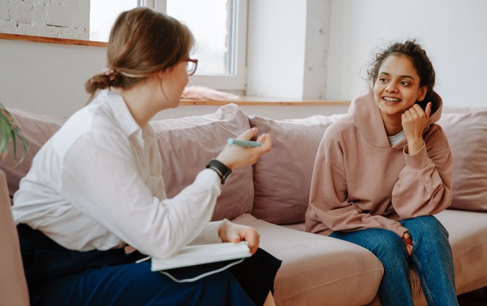 Two women sitting on a sofa talking to each other, one holding a notebook and pen