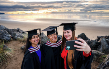 Three females dressed in graduation robes pose for a selfie on the beach 