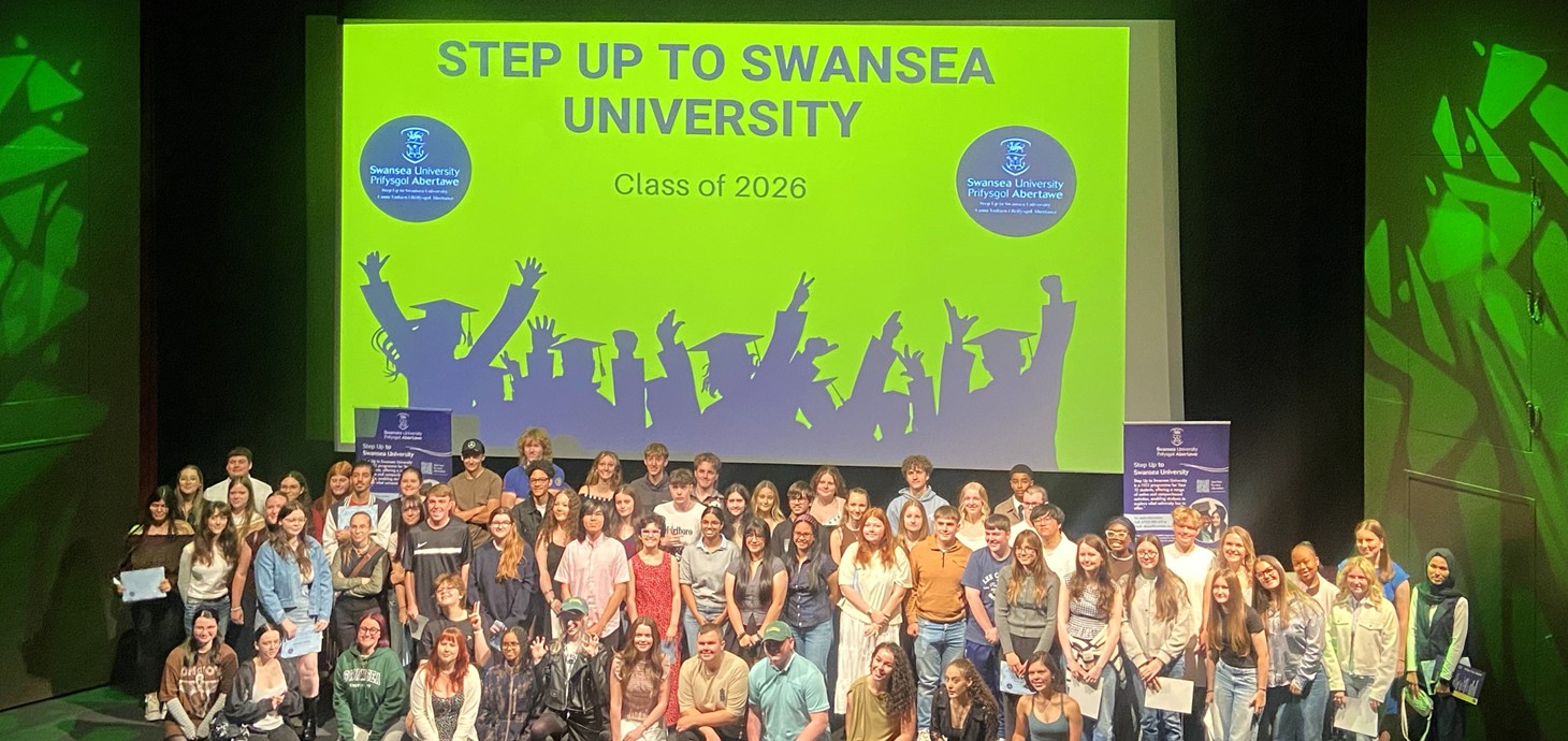 A large group of young people gathered on stage in front of a large green screen that reads 'Step Up to Swansea University' 