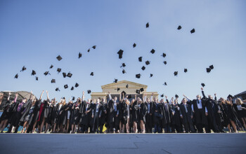 A large group of students in graduation robes throw their mortarboards in the air while standing in front of Swansea University’s Great Hall.