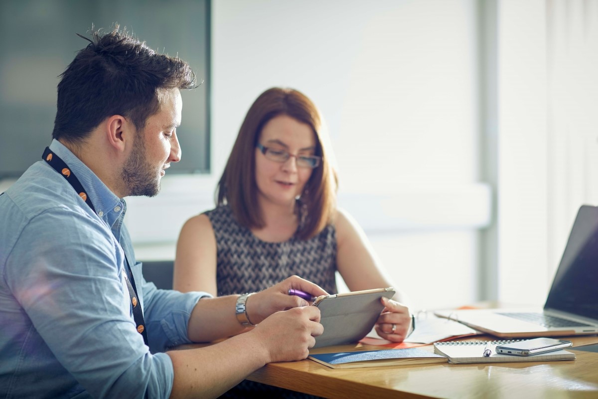 A male and female sitting at a desk and looking at an iPad.