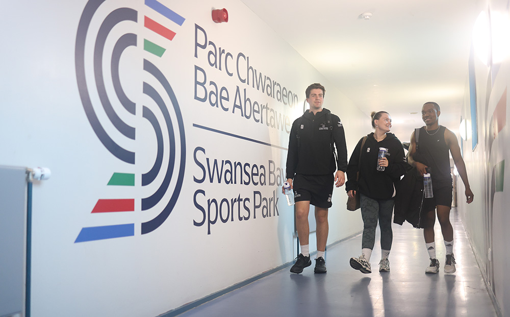 Three students wearing Swansea University Sport kit walking down the corridor to the Sports Hall and gym on Sketty Lane - a large Swansea Bay Sports Park logo is on the corridor wall