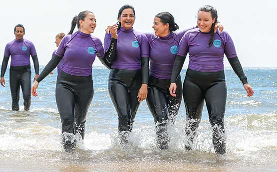 a group of female students in purple wetsuits standing in the shallow water at the beach with their arms around each other