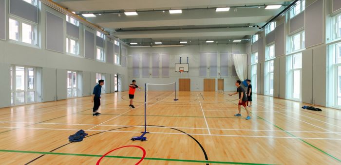 People playing badminton in a bright indoor sports hall with wooden courts